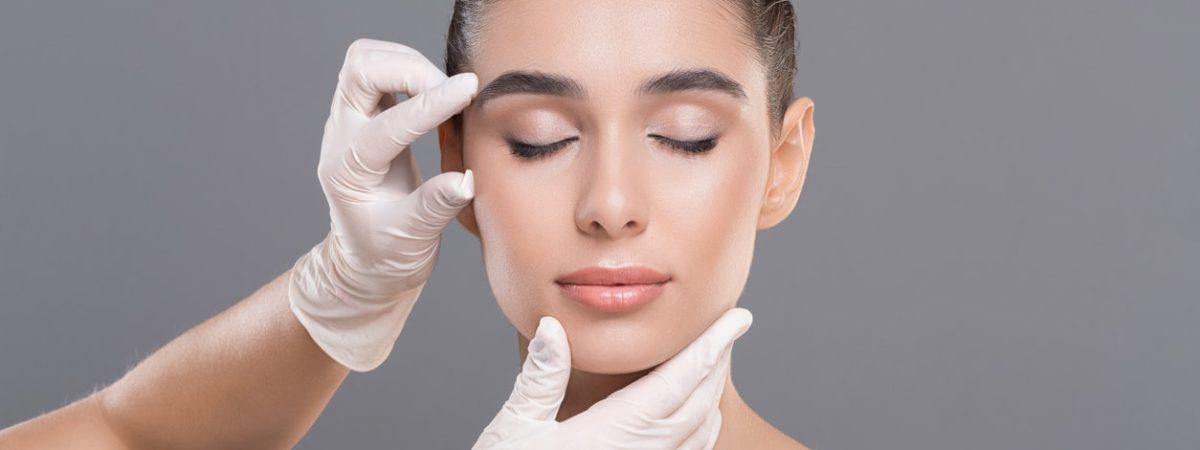 Dentist hands examining facial wrinkles on a young woman's face on a gray background.