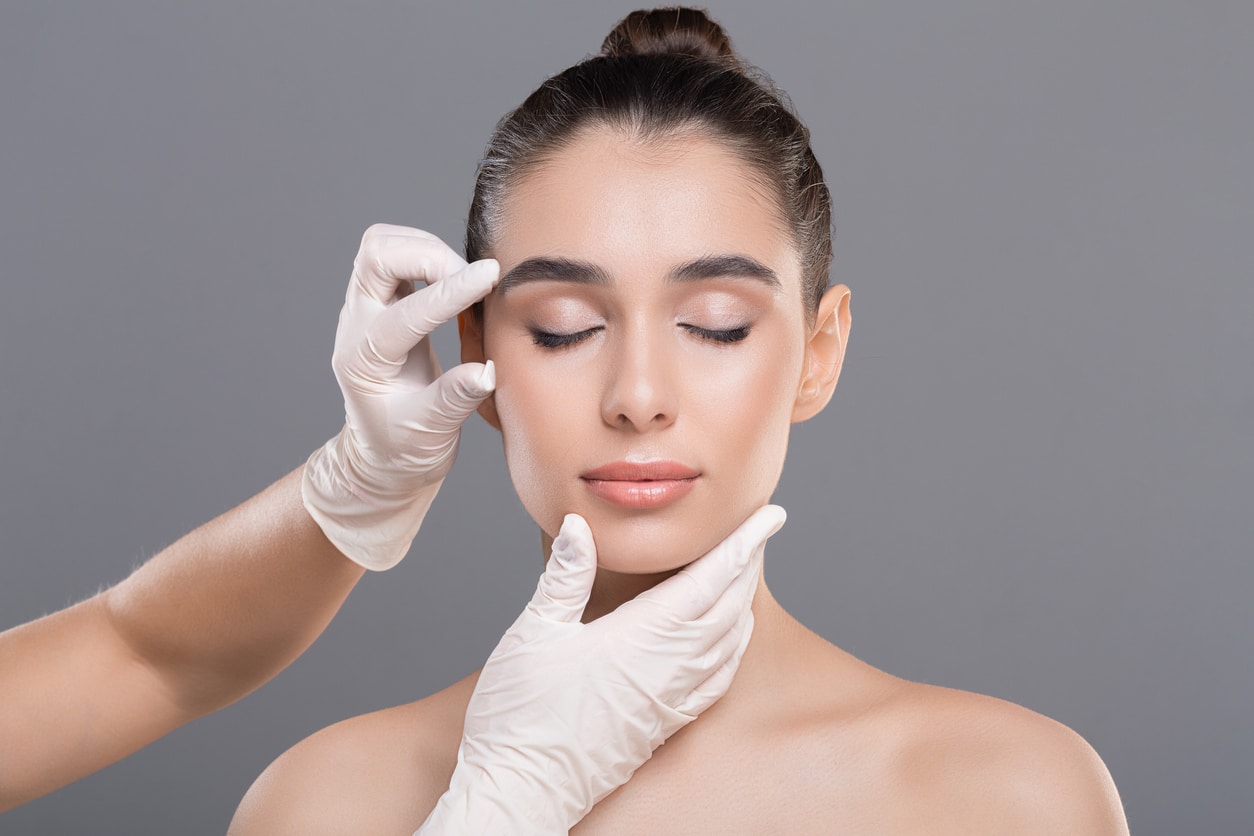 Dentist hands examining facial wrinkles on a young woman's face on a gray background.