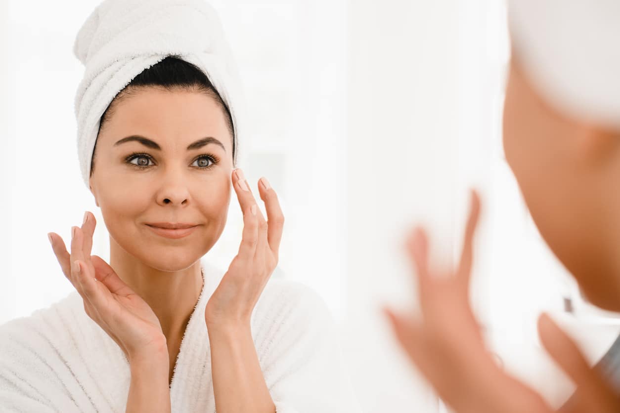 Woman in a spa robe and hair towel applying lotion after Botox in the mirror.