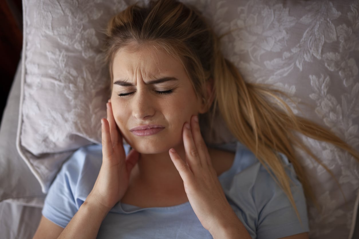 A woman with blonde hair and a blue shirt is in bed, grasping her jaw and experiencing TMJ pain.