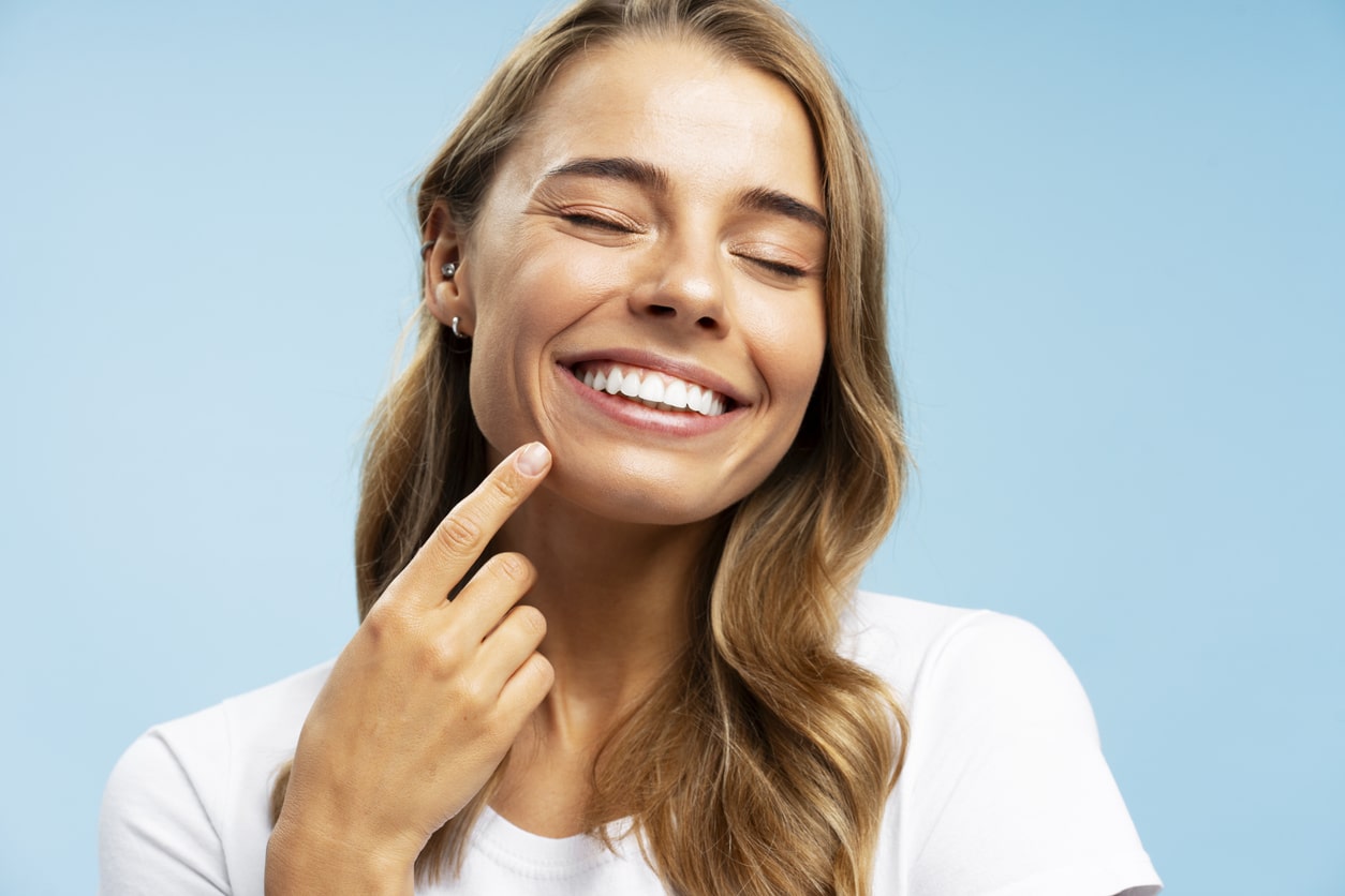 A woman with long, dirty-blonde hair points to her smile after following the essential steps to preventing tooth loss.