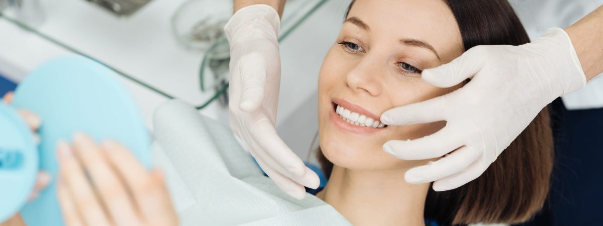 Dentist hands and happy patient looking in mirror after treatment A woman with shoulder-length brown hair sits in a dental chair and smiles after undergoing advanced cosmetic dental work.
