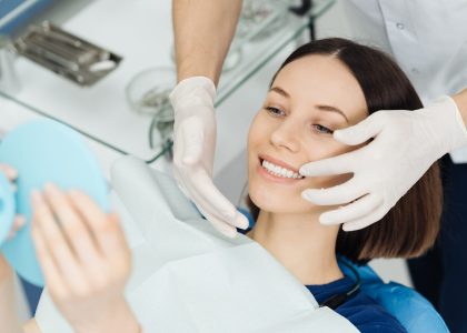 A woman with shoulder-length brown hair sits in a dental chair and smiles after undergoing advanced cosmetic dental work.