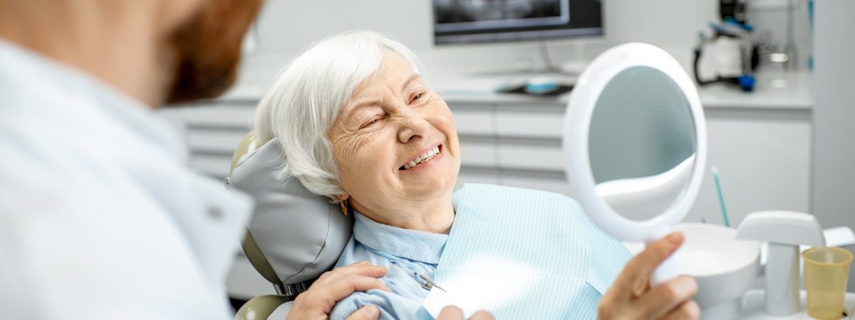 A smiling elderly woman sits in a dentist's chair, weighing her options between dentures and dental implants.