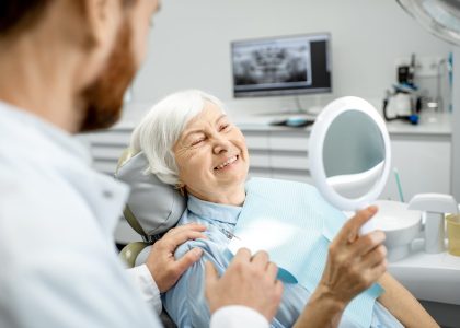 A smiling elderly woman sits in a dentist's chair, weighing her options between dentures and dental implants.