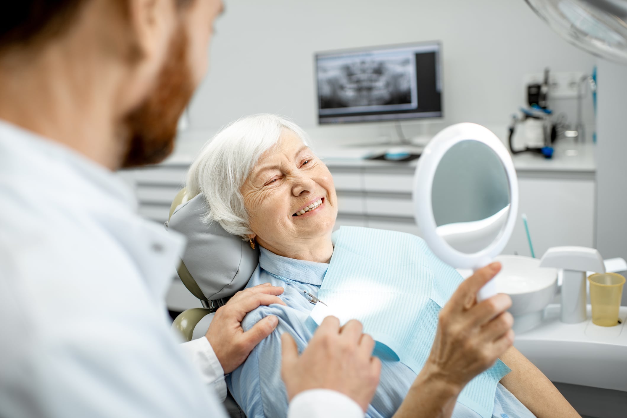 A smiling elderly woman sits in a dentist's chair, weighing her options between dentures and dental implants.