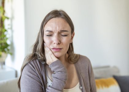 A woman sitting on a couch holds her jaw in pain and considers seeking professional TMJ treatment.