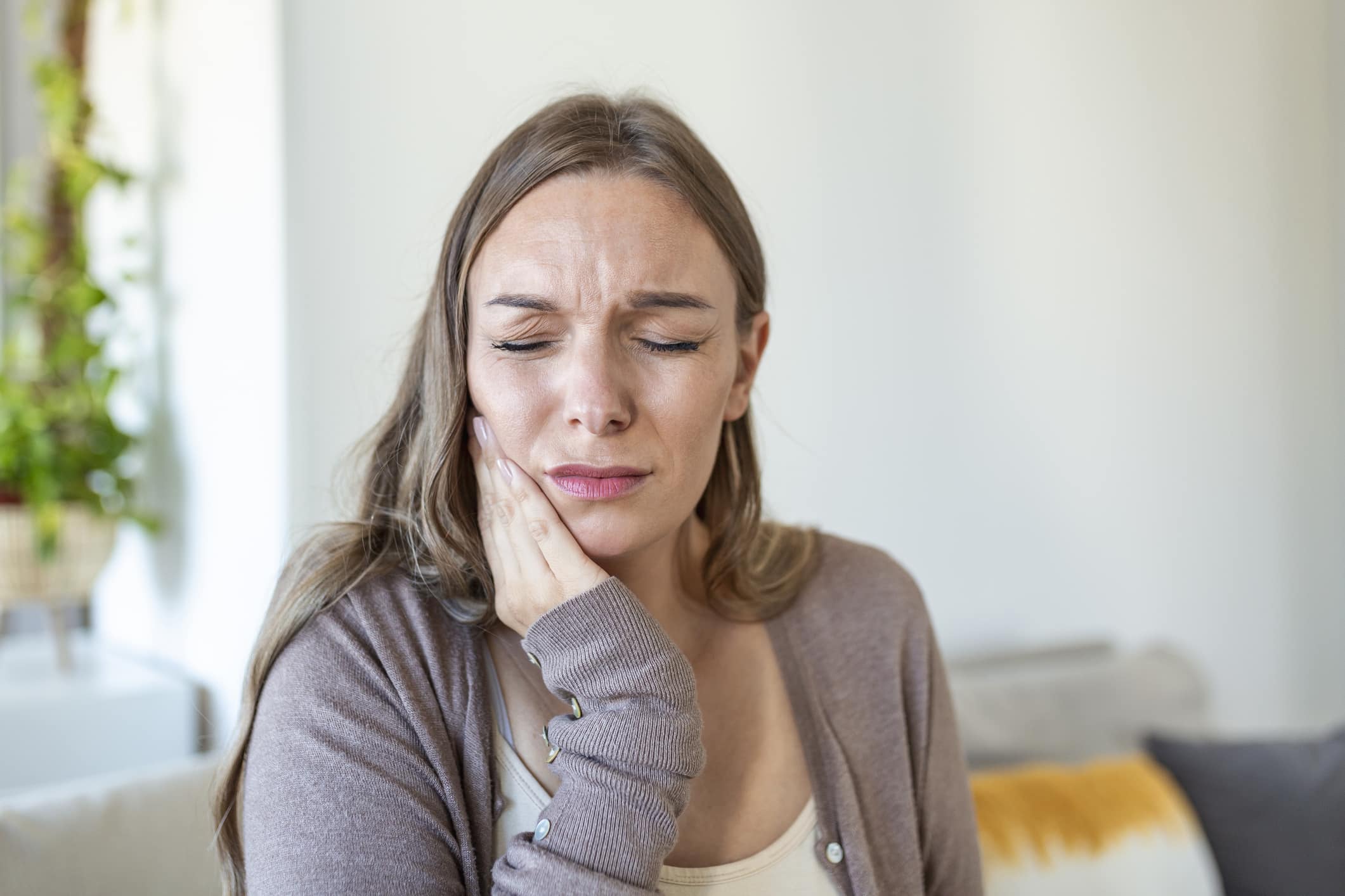 A woman sitting on a couch holds her jaw in pain and considers seeking professional TMJ treatment.